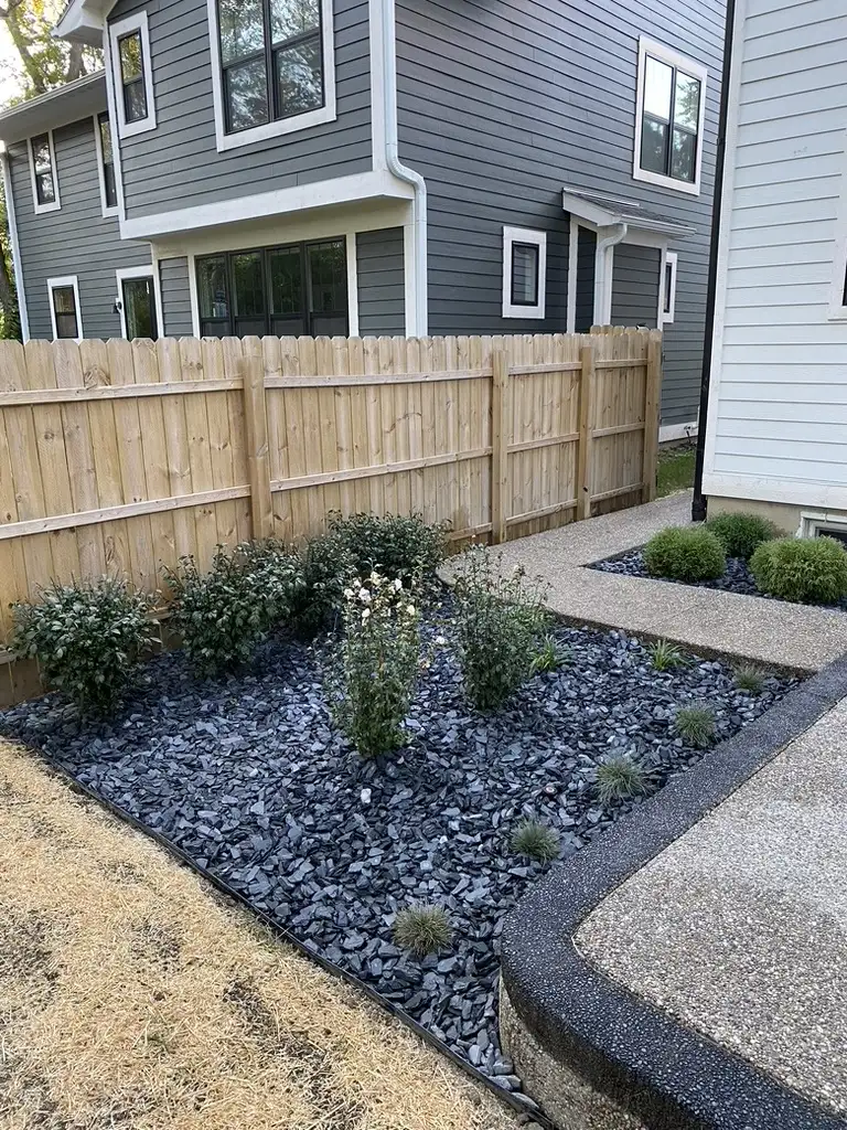 Landscaped side yard with a curved walkway, shrubs, ornamental grasses, and dark decorative stone