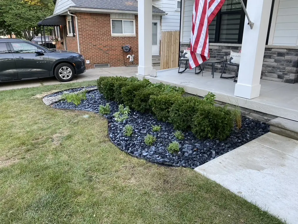 Curved front planting bed with evergreens, flowering plants, and dark decorative stone